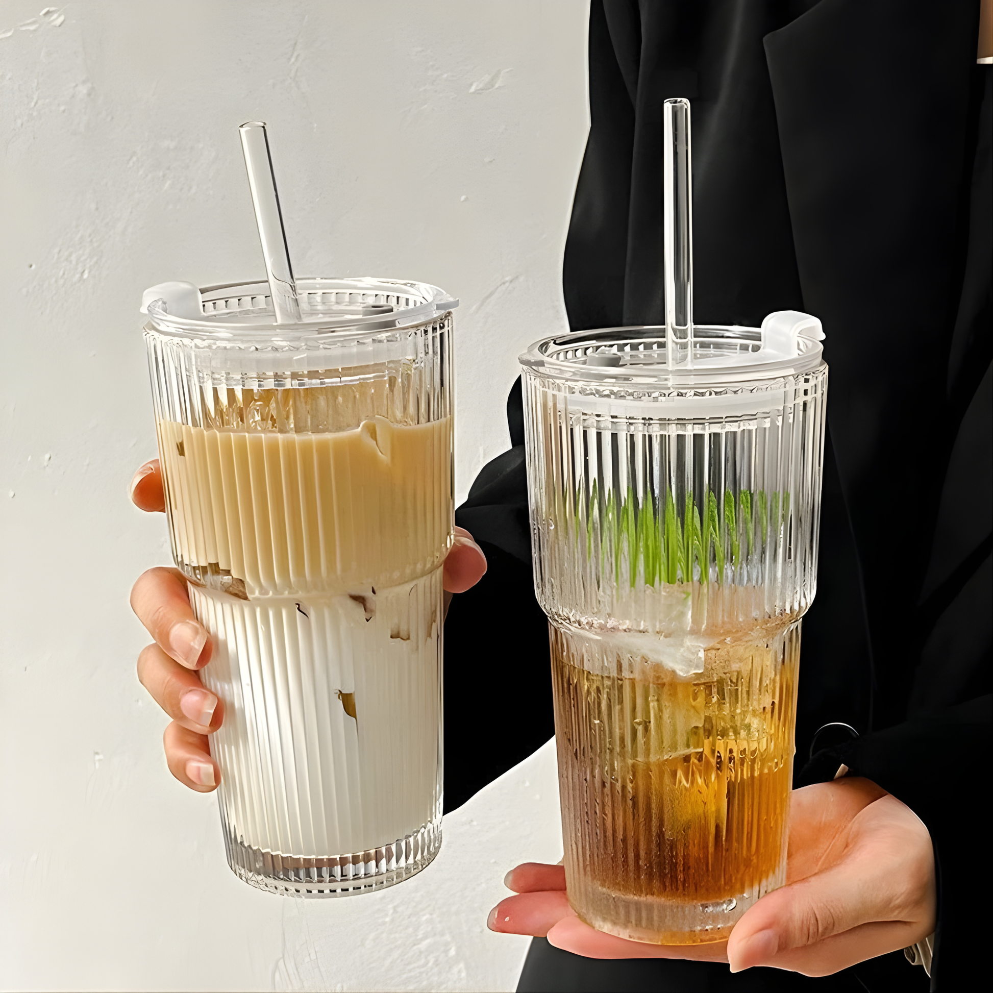 Deux grands verres striés de 600ml tenus en main, l'un contenant un café latte glacé et l'autre un thé froid, équipés de leur couvercle de voyage pour une dégustation nomade stylée.