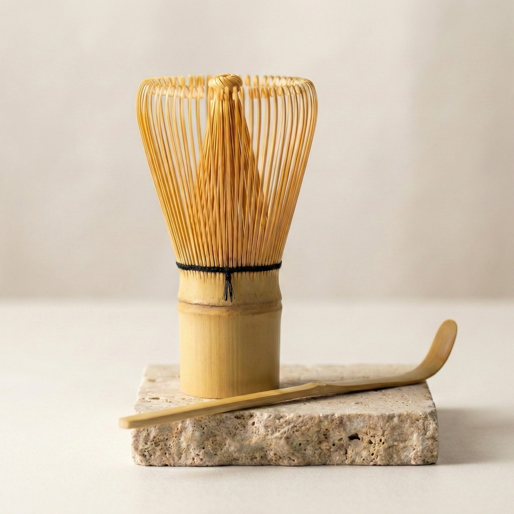 Photographie studio montrant un fouet à matcha en bambou doré (Chasen) avec sa ficelle noire, debout sur une dalle carrée de pierre travertin poreuse beige. Une cuillère à matcha traditionnelle en bambou (Chashaku) est posée en diagonale devant le fouet. L'arrière-plan est flou et clair avec un éclairage naturel doux.