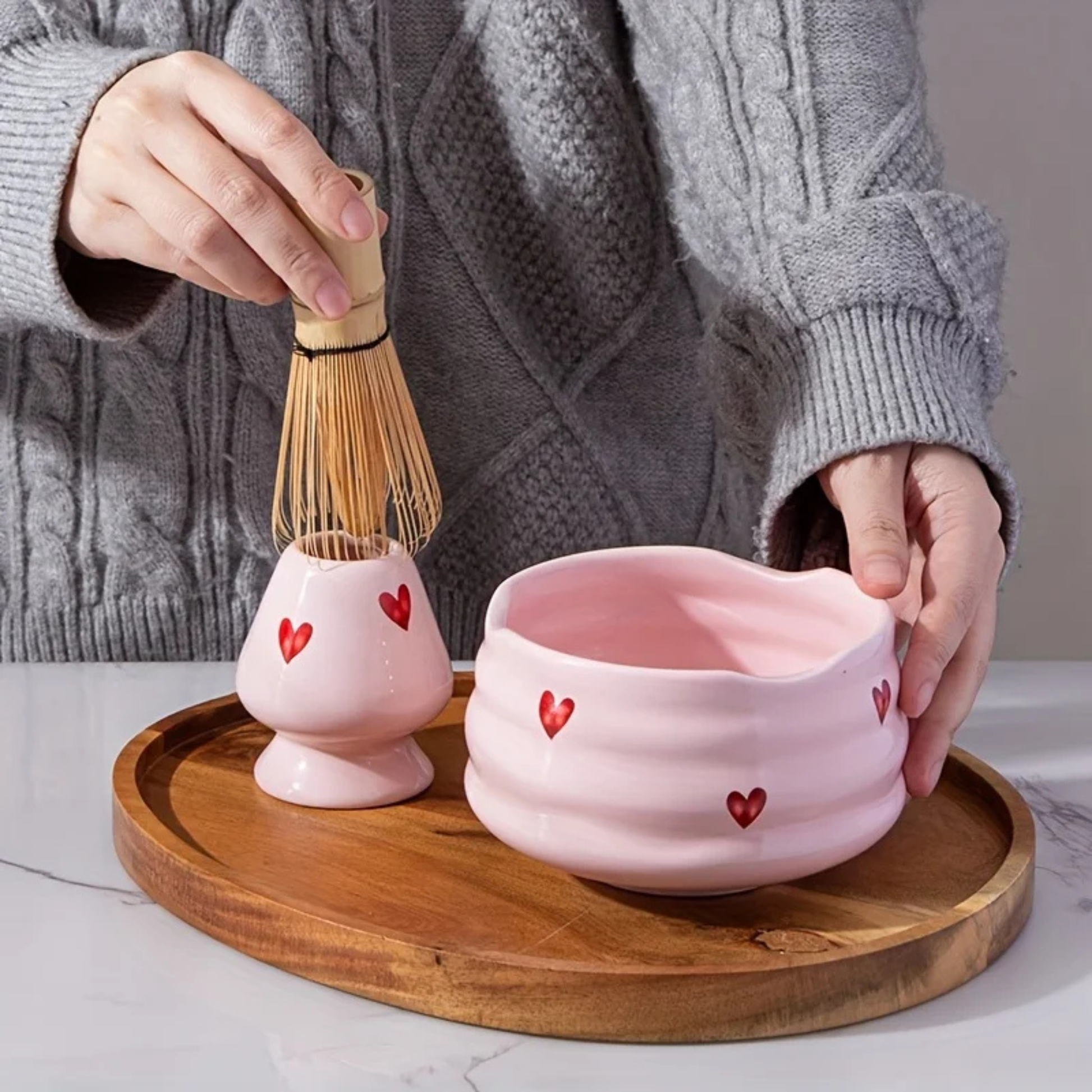 Mains préparant un thé matcha dans le bol Sakura rose, illustrant la taille généreuse du bol pour le fouettage et le design "Cœur" parfait pour offrir un moment de douceur.